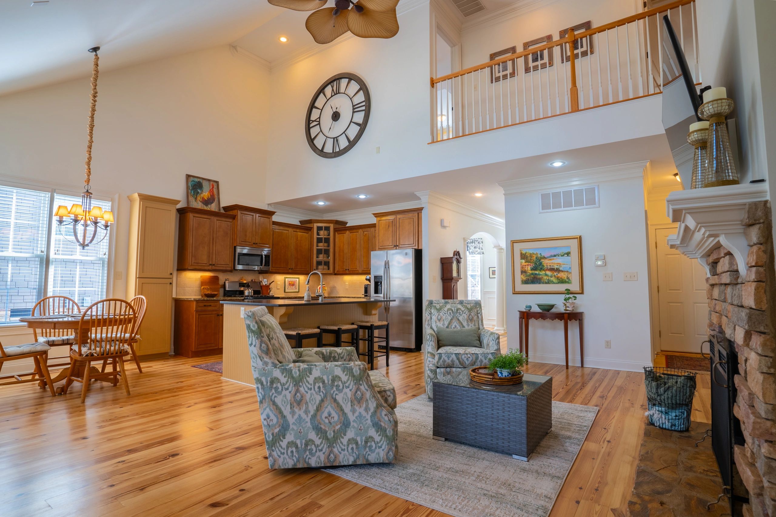 Open living room and kitchen with wood floors, a stone fireplace, and an upstairs balcony.