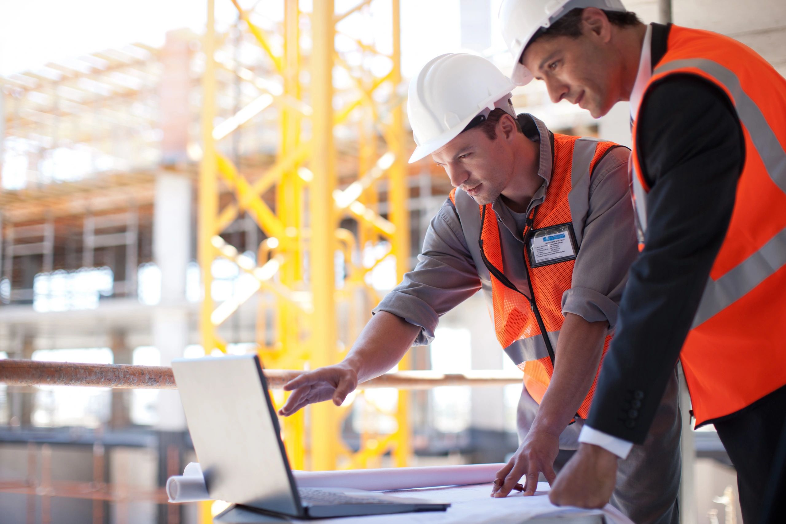 Two construction professionals in hard hats reviewing a laptop and plans at a jobsite.