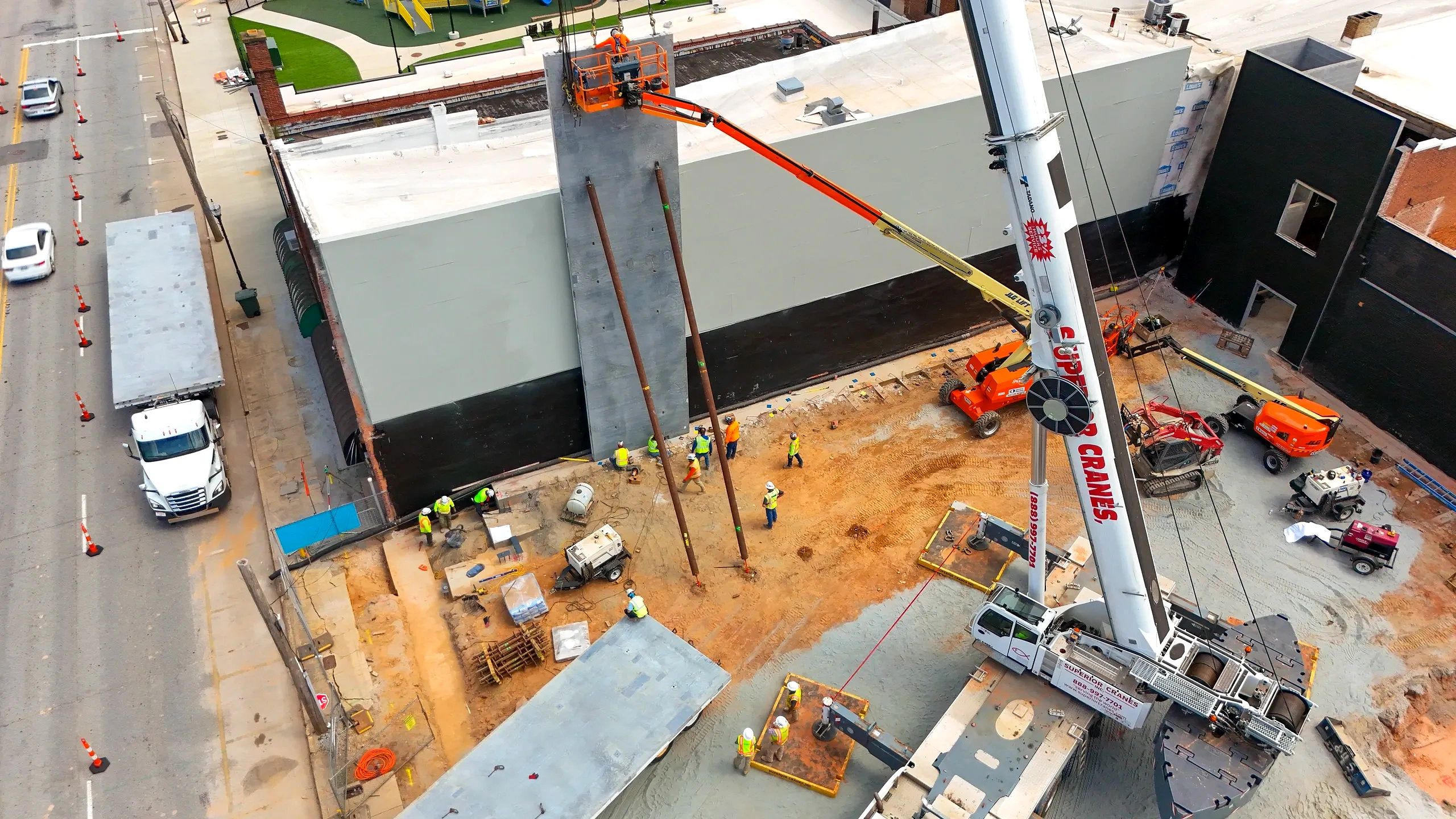 Aerial view of workers and a crane installing a concrete wall panel at a commercial construction site.