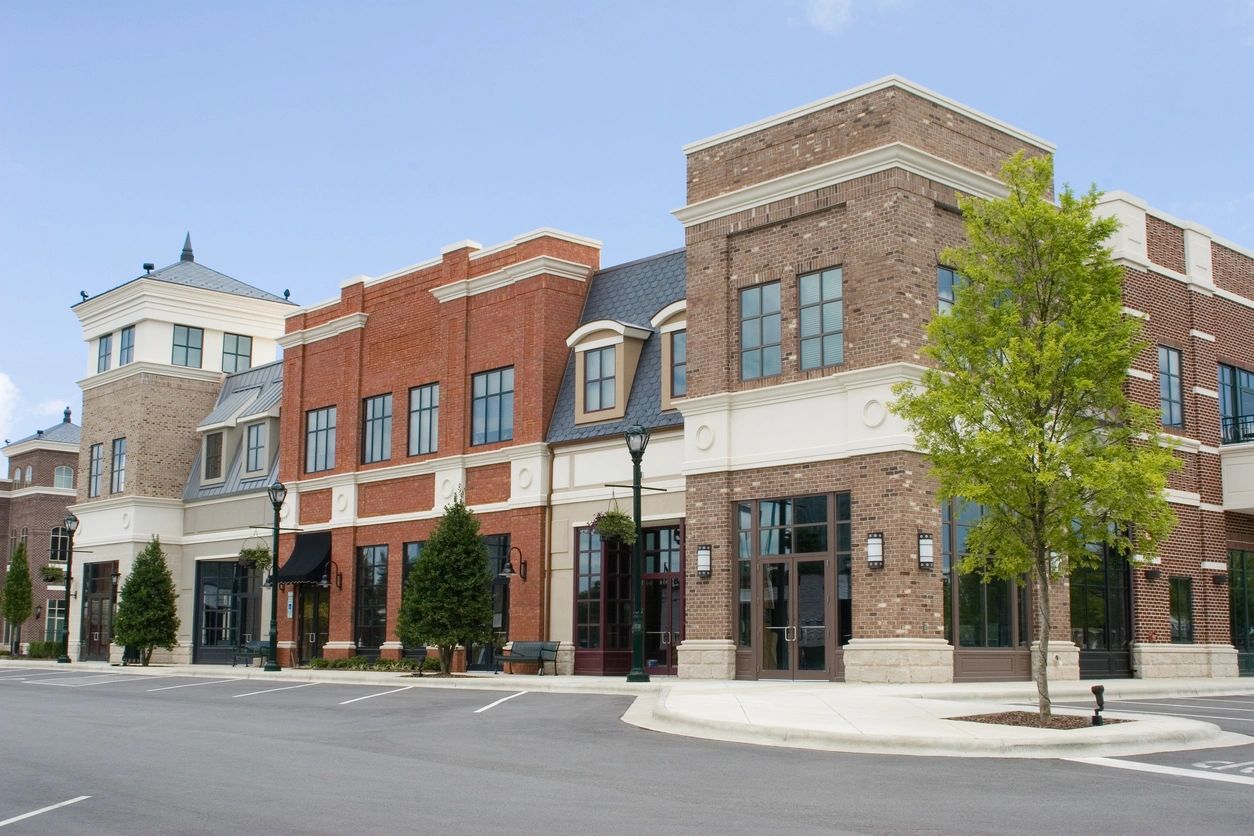 Brick commercial storefronts with sidewalks, street trees, and an empty parking area.