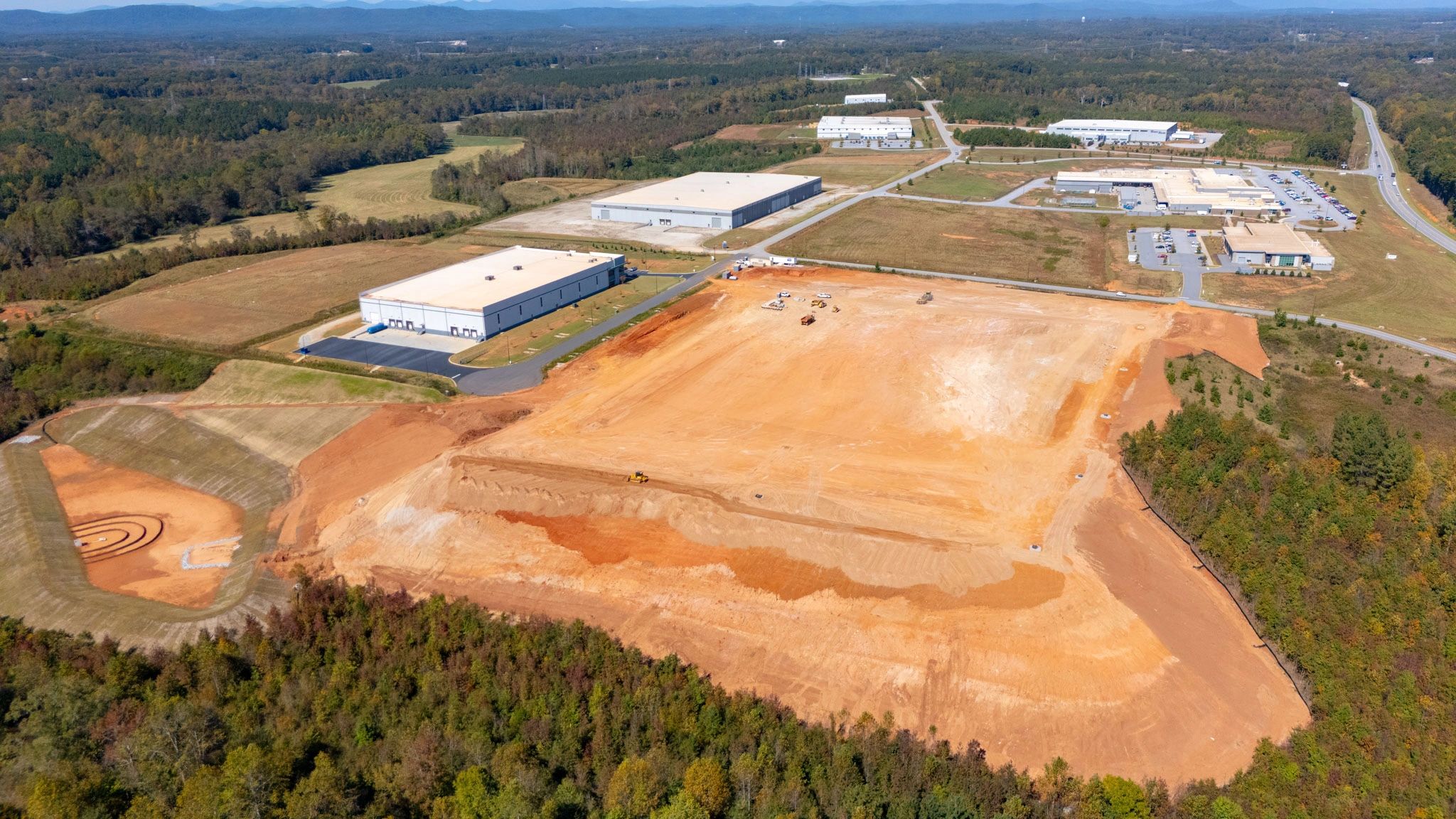 Wide aerial view of a graded industrial construction site, stormwater basin, and nearby warehouses.